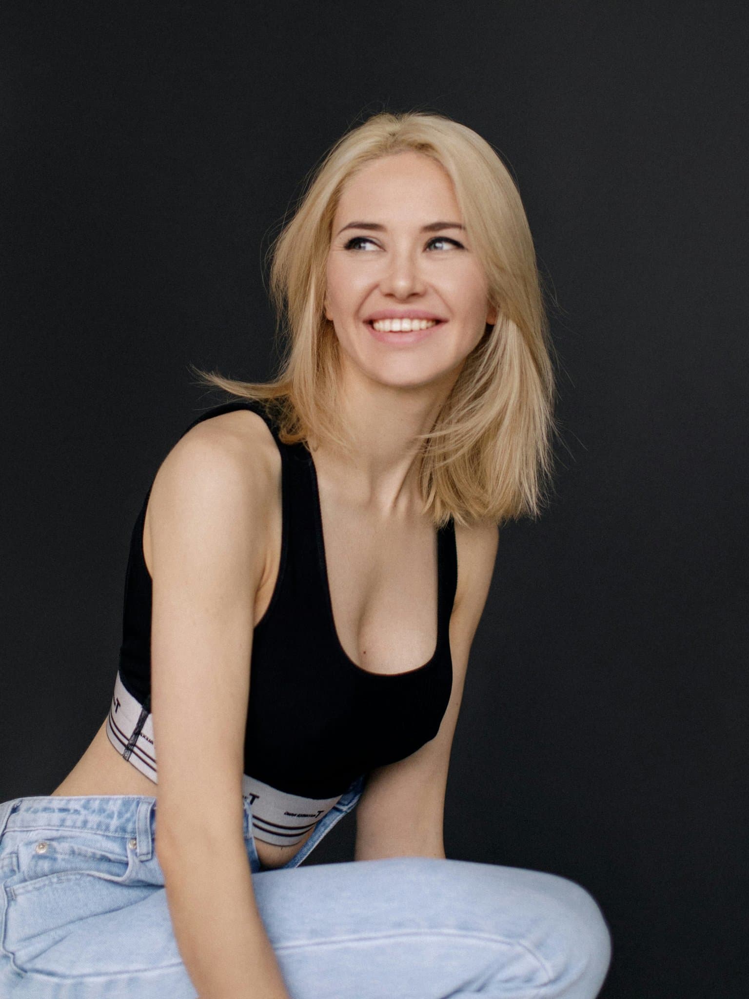 Casual portrait with blonde hair, black crop top, light blue jeans, smiling against dark background.