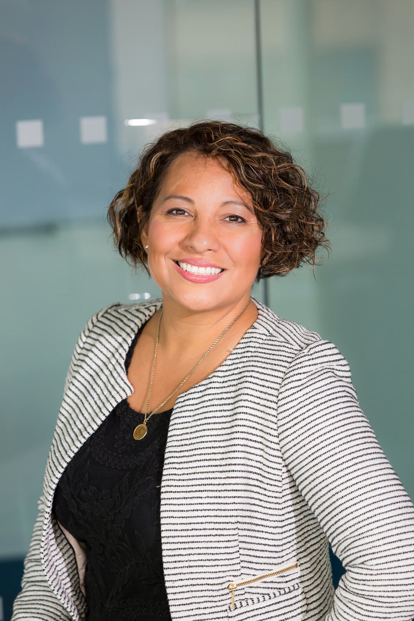 Professional portrait with short curly hair, striped blazer, black top, warm smile in office setting.