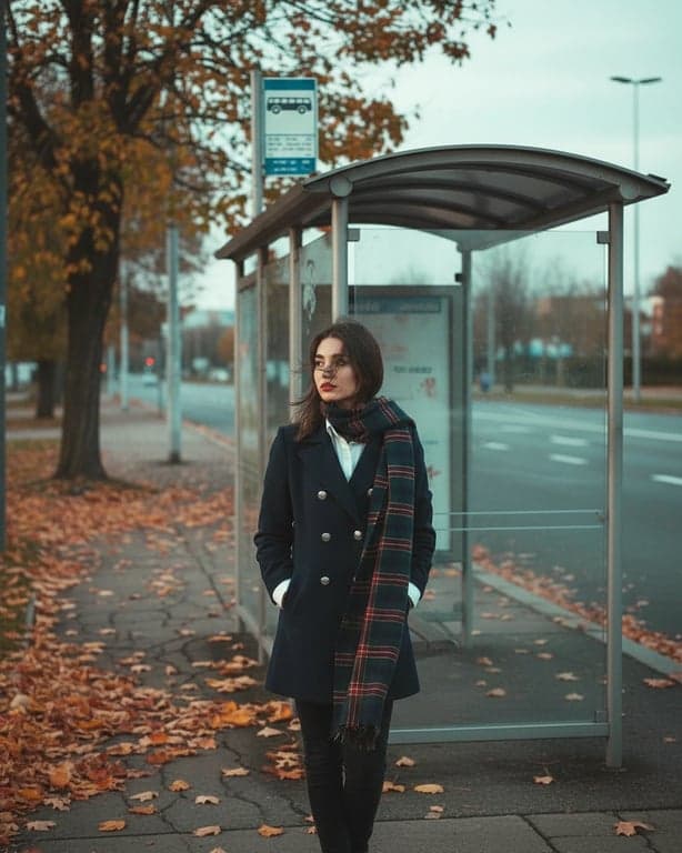 a portrait of a student transformed into a winter uniform with a coat and scarf, waiting at a bus stop with autumn leaves