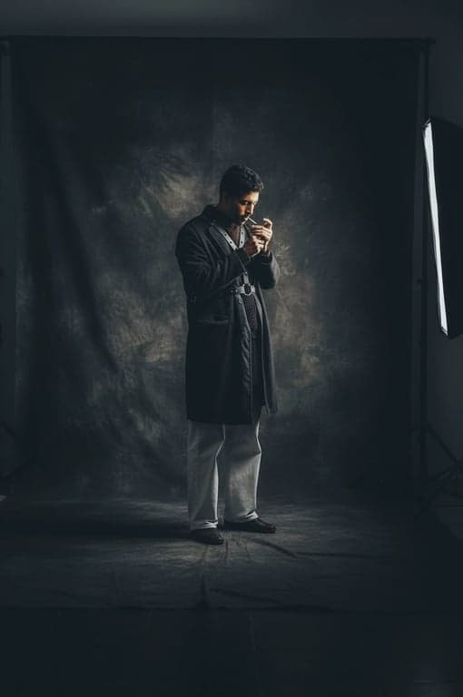 a portrait of a man in a leather harness and mesh top outfit, standing in a moody studio with dramatic side lighting