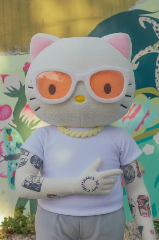 Convert the original person into a Hello Kitty parallel world plush character with a cat face. Preserve pose and outfit shape, render the character as a soft plush toy with rounded forms, fuzzy fabric texture, stitched-like edges, and gentle pastel colors, styled like official Sanrio plush merchandise.